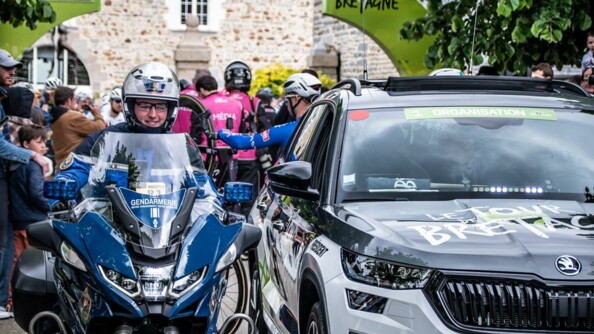 Au premier plan, un motocycliste de la garde républicaine à côté d'un véhicule de course. Au second plan, des coureurs et des spectateurs.