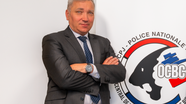 Le colonel Hubert Percie du Sert, commandant de l'Office central de lutte contre le trafic des biens culturels, en costume gris, à côté du logo de l'OCBC.