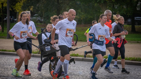Des coureurs, vêtus d'un T-shirt blanc avec un numéro sur le ventre, courent sur une avenue pavée, en poussant le fauteuil roulant d'une jeune femme blonde, portant des tresses. On aperçoit des arbres aux feuilles vertes en arrière plan.