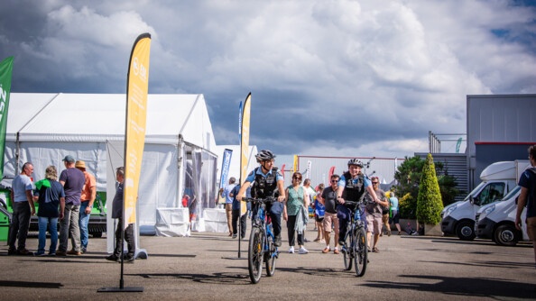 Deux gendarmes à vélo en patrouille autour des abords d'un salon. De part et d'autres des visiteurs, quelques camions et tentes d'exposition.