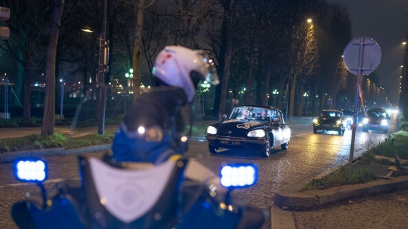 Un gendarme en moto de nuit devant des véhicules de collection