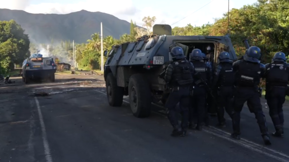 Deux véhicules blindés avancent vers un barrage érigé sur une route en Nouvelle-Calédonie. suivis par des gendarmes marchant en ordre serré à l'arrière du second véhicule