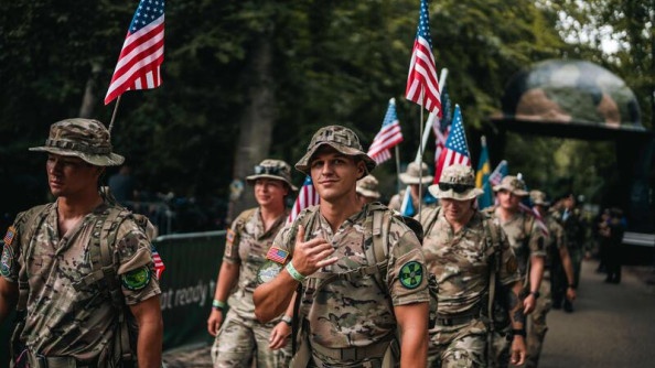 Un groupe de militaires, en treillis kaki, marchent ensemble, face à l'objectif. Plusiers d'entre eux portent un drapeau américain, accroché à leur sac à dos.