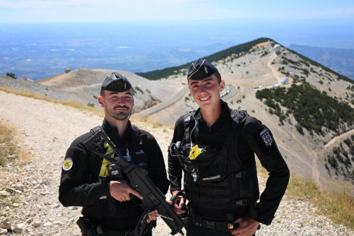 Deux jeunes gendarmes, vêtus d'une tenue noire, se tiennent debout, côte à côte, sur les hauteurs du Mont Ventoux. Le temps estc lair et ensolleilé.