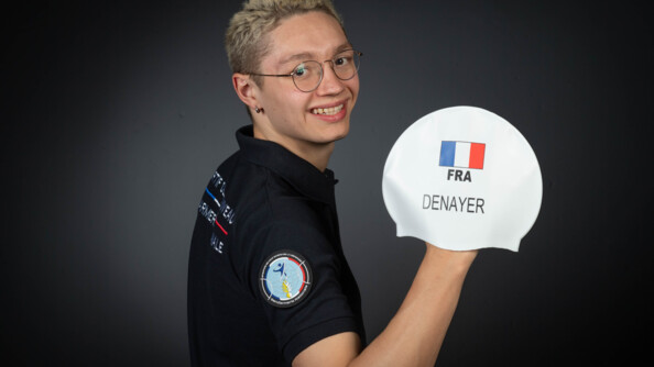 Un jeune homme blond à lunette, portant un polo noir et arborant l'écusson rond des sportifs de haut niveau de la Défense-gendarmerie, montre de sa main droite son bonnet de piscine siglé FRA avec un drpeau bleu-blanc-rouge et de son nom, Denayer