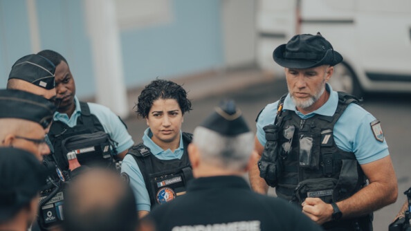 Une femme gendarme, brune aux cheveux courts ondulés, et vêtue d'un gilet pare-balles portant l'inscription "Gendarmerie", se tient debout au milieu d'hommes.