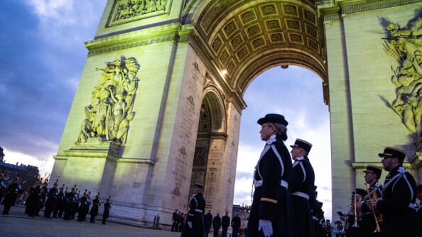 Vue en contre-plong&eacute;e de trois-quart sur l'Arc de Triomphe &eacute;clair&eacute;, tandis que la nuit tombe. Au premier rang, une rang&eacute;e de gendarmes au garde-&agrave;-vous. Derri&egrave;re eux des musiciens (trompettistes). Au second plan, les troupes au garde-&agrave;-vous.