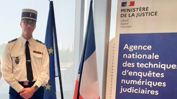 Un capitaine de gendarmerie, debout à gauche de l'image, en chemise blanche et pantalon bleu, coiffé de son képi, les mains croisées devant lui. A sa droite, les drapeaux de l'Union uropéenne et de la France, puis un kakémono de l'Agence nationale des techniques d'enquêtes numériques judiciaires
