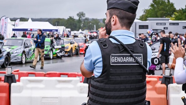 Un gendarme de dos derrière des barrières en plastiques rouge et blanche,limitant l'accès aux voitures de courses de diverses couleurs et ay public et coureurs sur place