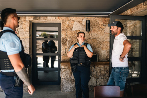 Deux gendarmes discutent avec un homme vêtu d'un jean, d'un t-shirt blanc et d'une casquette noire, se tenant à l'entrée d'une habitation
