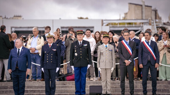 Des militaires hauts gradés, dont le directeur général de la gendarmerie nationale, se tiennent debout, aux côtés d'élus portant sur la poitrine l'écharpe tricolore. Derrière eux est installé un cordon de sécurité de couleur blanche, derrière lequel se trouve une foule.