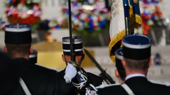 Un militaire vêtu d'une tenue sombre et d'un képi noir gansé de blanc, tient devant lui son épée pointée vers le ciel. Sa main tenant l'épée est poitionnée au niveau de son visage. Des militaires, que l'on aperçoit de dos, lui font face. Derrière, en aperçoit les gerbes de fleurs dans des tons bleu, blanc, rouge.