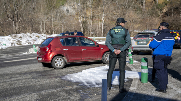 a droite de la photo, un gendarme et un policier espagnol tenant ses mains derrière le dos, devant un contrôle routier où deux gendarmes controlent un véhicule rouge au bord de la route de montagne