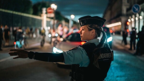 Une jeune femme gendarme se tenant debout, dans une rue de Paris, guide les voitures. Elle est équipée d'un gilet pare-balles portant dans le dos l'inscription "Gendarmerie".La scène se déroule la nuit.
